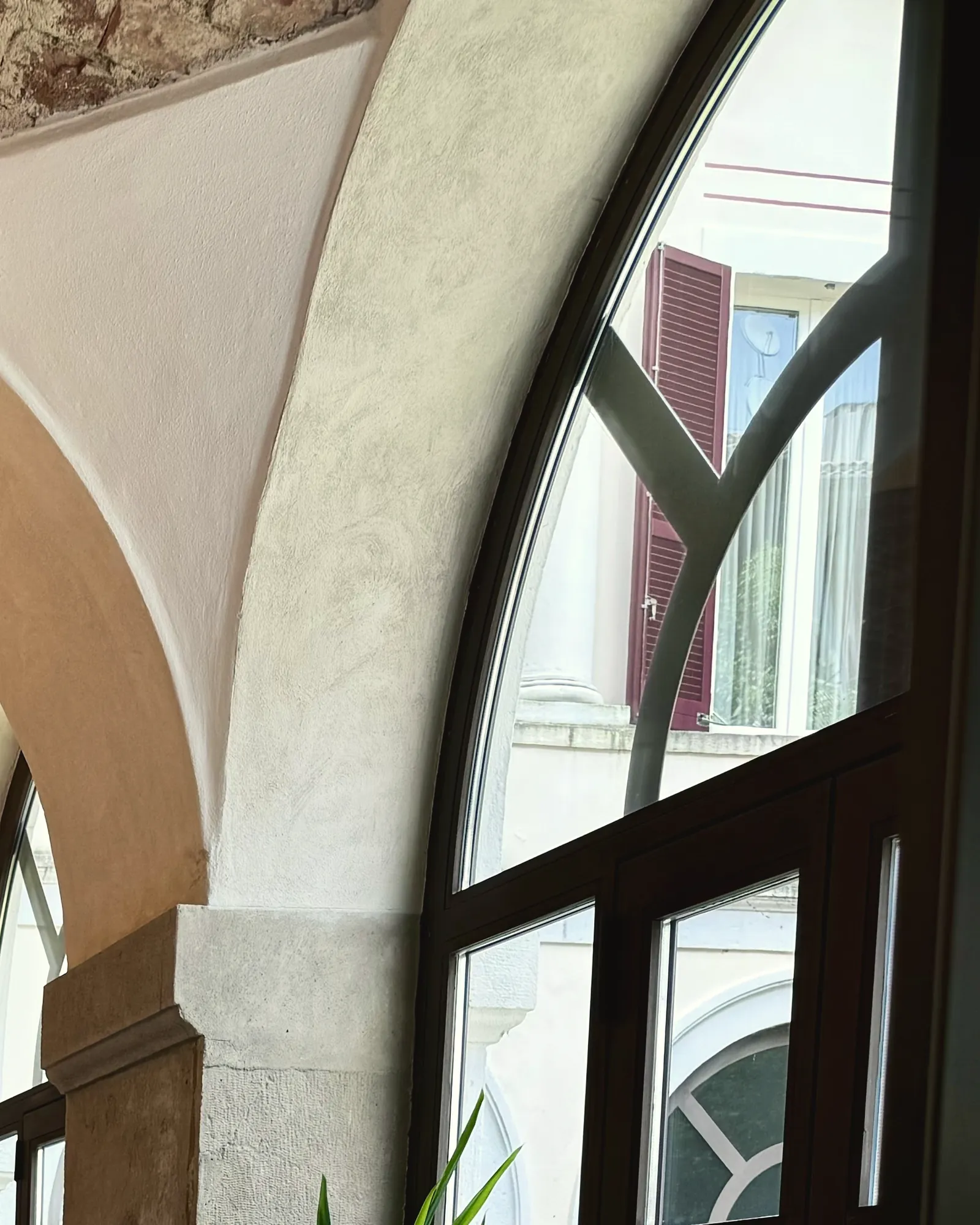 Una delle grandi finestre ad arco dello studio di via Donizetti 4, coi telai scuri, accanto a una colonna chiara: fuori, una facciata con le persiane rosse.