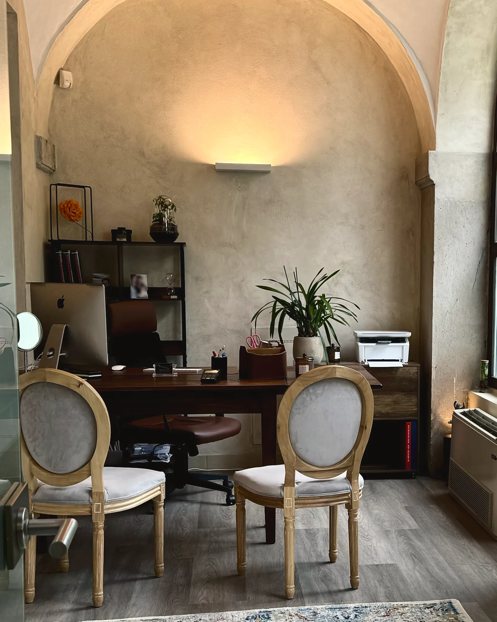 La scrivania dello studio di via Donizetti 4 e due sedie imbottite in legno chiaro, sotto la volta, con una lampada calda sulla parete.
