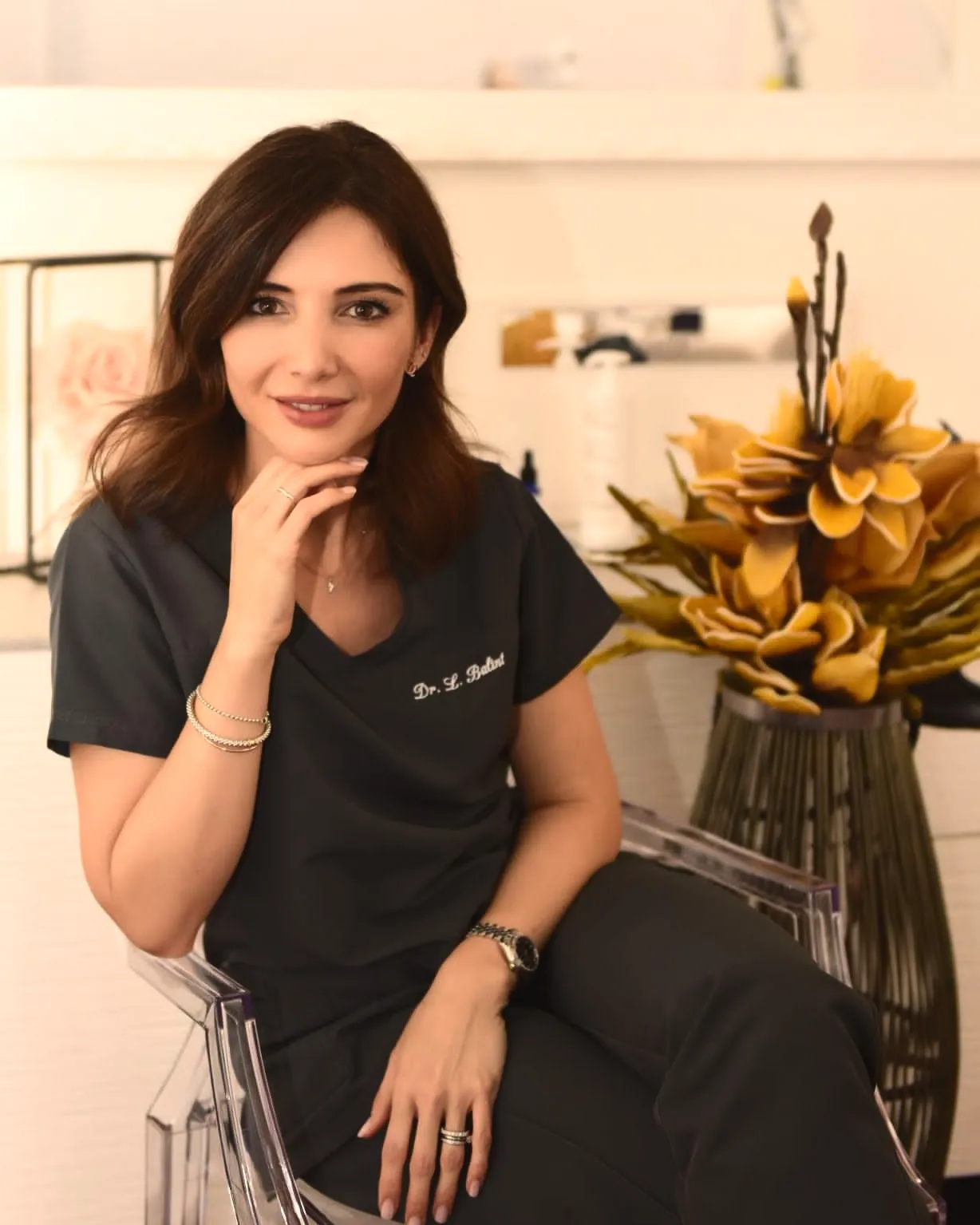 La Dott.ssa Laura Balint seduta in studio, in divisa scura col suo nome ricamato e una mano al mento; accanto un vaso di fiori dorati.